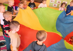 dzieci na dywanie stoją z kolorową chustą animacyjną trzymają jej brzegi, na niej leża małe maskotki